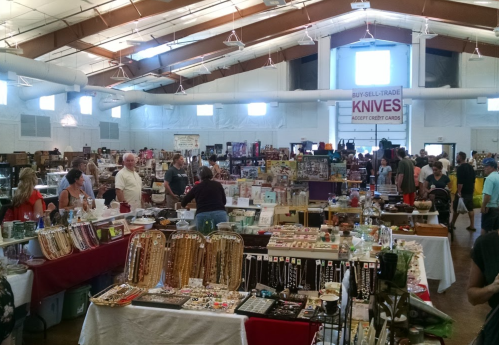 A bustling indoor market with various vendors selling items, including jewelry and collectibles, and many shoppers browsing.