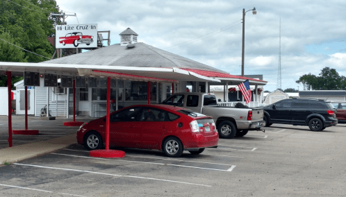 A diner with a red car and a truck parked outside, featuring a sign that reads "Hi-Lite Cruz-In."