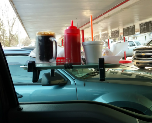 A car tray holds a glass of soda, ketchup, and a takeout container, resting on the window of a parked vehicle.