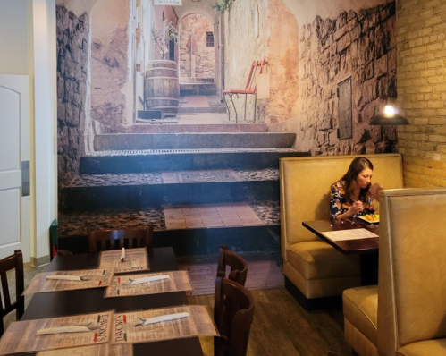 A woman sits at a table in a restaurant with a mural of a cobblestone alleyway on the wall behind her.