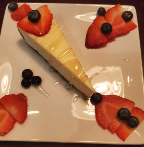 A slice of cheesecake topped with strawberries and blueberries, drizzled with syrup on a square plate.