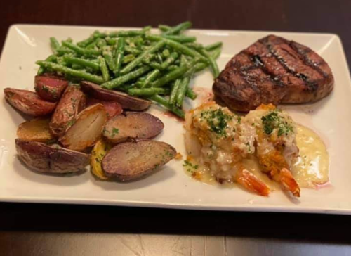 A plate with grilled steak, green beans, roasted potatoes, and shrimp with a creamy topping.
