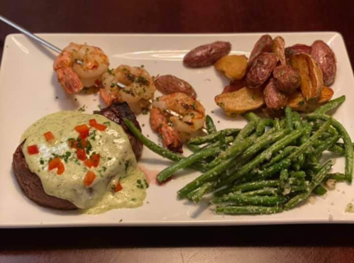A plate featuring a steak with sauce, shrimp, roasted potatoes, and green beans.