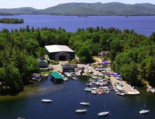 Aerial view of a marina with boats, surrounded by lush greenery and mountains, overlooking a calm lake.
