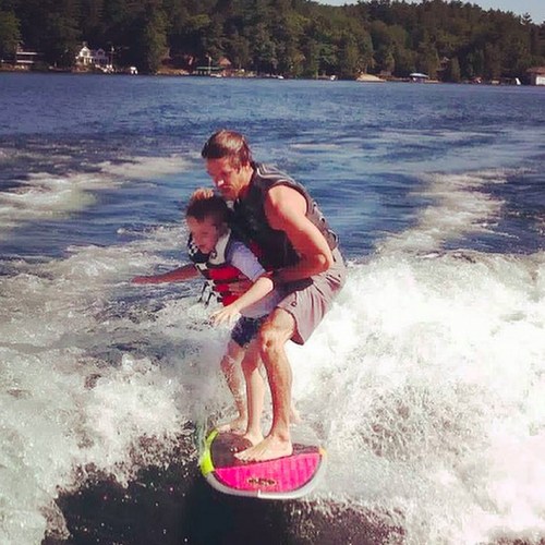 A man and a child are surfing together on a colorful board, riding a wave on a sunny day.