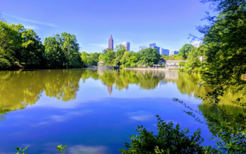 A serene lake reflects city skyscrapers and lush greenery under a clear blue sky.