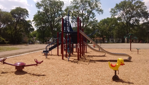 A playground with a slide, climbing structure, and a yellow duck spring rider, surrounded by wood chips and trees.