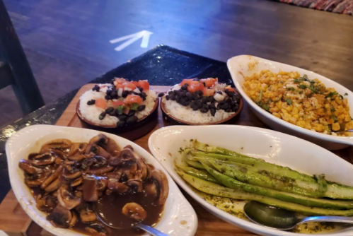 A wooden platter with rice topped with black beans and tomatoes, corn salad, sautéed mushrooms, and grilled asparagus.