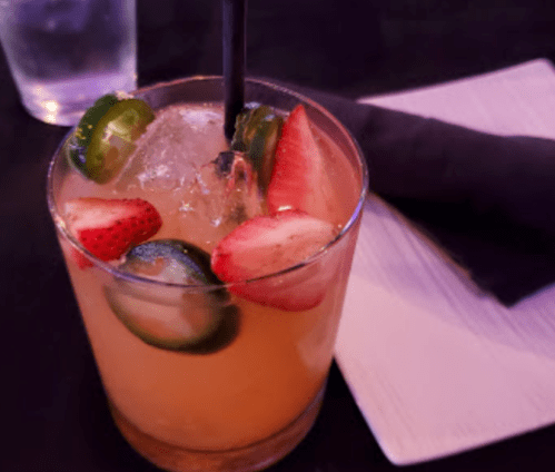 A refreshing cocktail with ice, strawberries, and cucumber slices, served in a glass with a straw on a dark table.