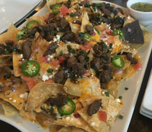A plate of nachos topped with cheese, beef, jalapeños, tomatoes, and herbs, served with a side of green salsa.