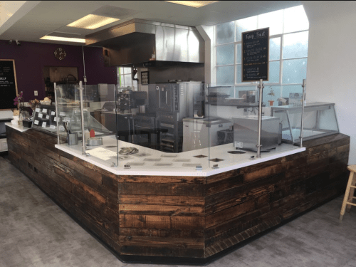 A modern café counter with a wooden finish, glass barriers, and kitchen equipment visible in a bright, open space.