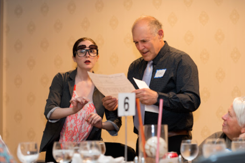 A woman in glasses and a man in a tie discuss a document at a dinner table, with guests seated around them.