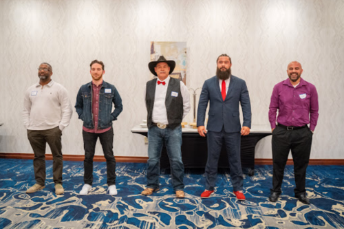 Five men stand in a row, each dressed in different styles, in a well-lit indoor setting with a patterned carpet.