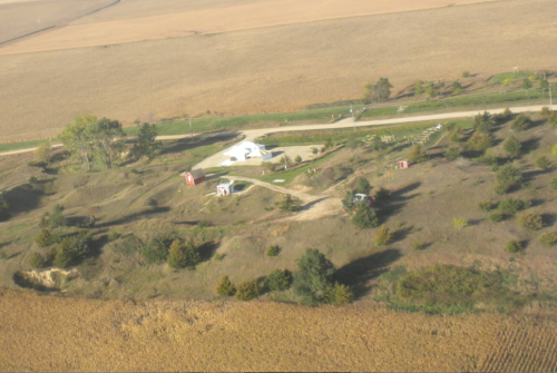 Aerial view of a rural landscape with fields, a road, and small structures among trees and shrubs.