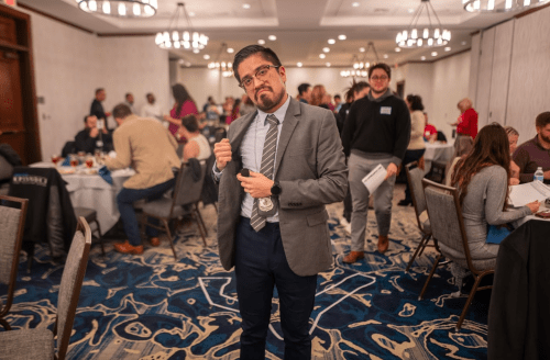 A man in a suit stands confidently in a busy event space with people mingling and tables set up in the background.