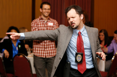 A man in a suit and badge dramatically points while dancing at a social event, with others laughing in the background.