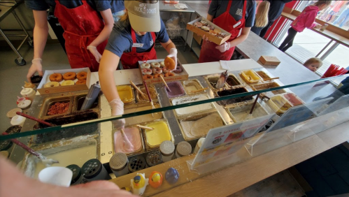 A colorful ice cream shop counter with staff serving ice cream and toppings, while a child watches from the side.