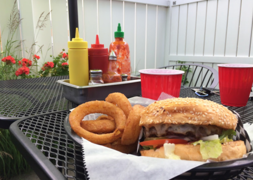 A sesame seed burger with lettuce and tomato, served with onion rings, condiments, and drinks on a patio table.