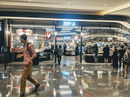 A bustling food court with various eateries, featuring neon signs and people walking through a shiny, tiled space.