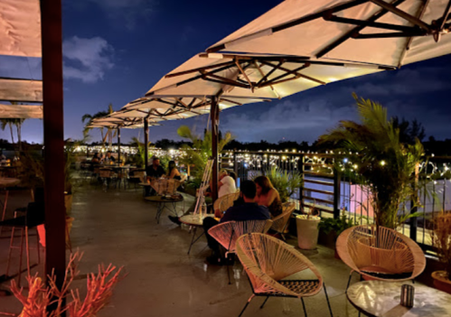 A cozy outdoor restaurant at night, with string lights, tables, and people enjoying the view by the water.