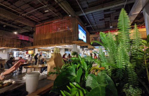 A vibrant indoor market with greenery, wooden structures, and people dining in a lively atmosphere.