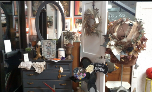 A cozy vintage shop display featuring a mirror, wreaths, and various decorative items on a dresser.