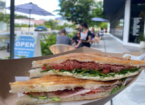 A close-up of a sandwich with layers of meat, cheese, and greens, held in a hand outside a café with patrons in the background.