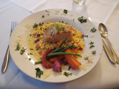 A plate of fish on a bed of creamy grains, garnished with green beans, carrots, and red peppers, with herbs sprinkled on top.