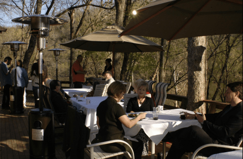A group of people dining outdoors on a deck, surrounded by trees and enjoying a sunny day.