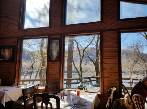 Cozy restaurant interior with wooden walls, large windows showing a scenic view of trees and a river outside.