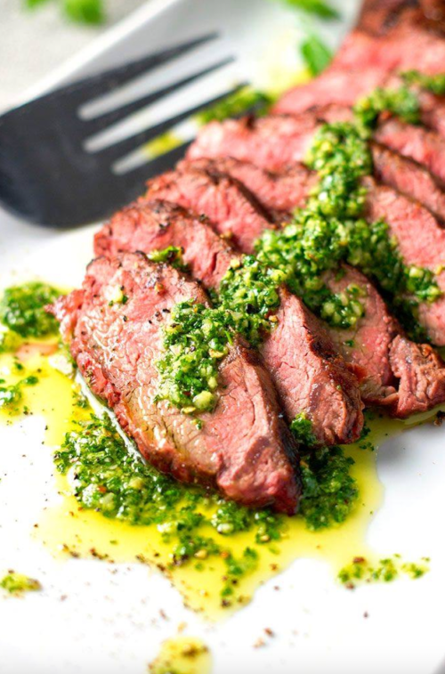 Sliced steak drizzled with green sauce, garnished with herbs, on a white plate with a spatula in the background.