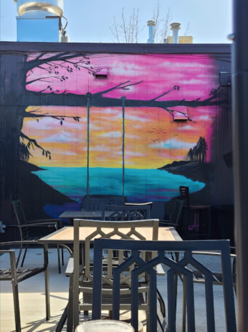 A vibrant mural of a sunset over water with swings, framed by trees, viewed from an outdoor seating area.