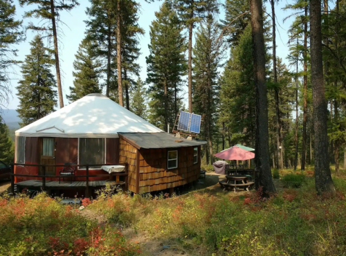 A cozy yurt surrounded by tall trees, with a solar panel on the roof and a picnic area nearby.