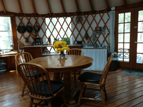 Cozy yurt interior with a wooden table, chairs, and a vase of yellow flowers, surrounded by rustic decor and natural light.