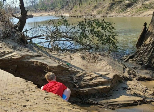 A child in a red shirt explores a sandy riverbank with trees and water in the background on a sunny day.