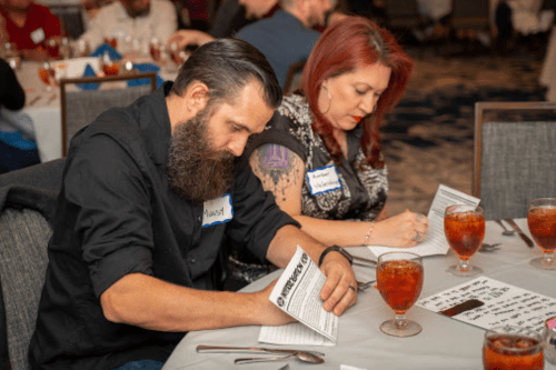 Two people seated at a table, writing on papers, with drinks in front of them at a social event.