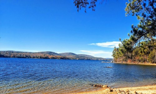 A serene lake surrounded by hills under a clear blue sky, with a sandy shore and scattered rocks.