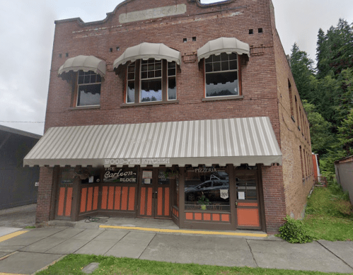 Historic brick building with large windows, striped awning, and signage for a pizzeria, surrounded by trees.