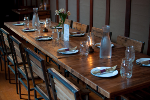 A long wooden dining table set with plates, glasses, and flowers, surrounded by rustic chairs in a cozy setting.