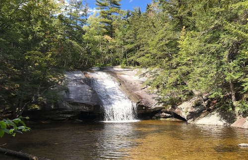 A serene waterfall cascades over smooth rocks into a clear pool, surrounded by lush green trees and a blue sky.
