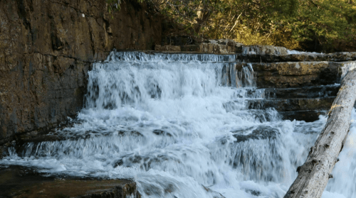 A cascading waterfall flows over rocky steps, surrounded by lush greenery and natural textures.