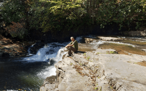 A person sitting on a rocky ledge by a flowing river with a waterfall in a lush, green forest setting.