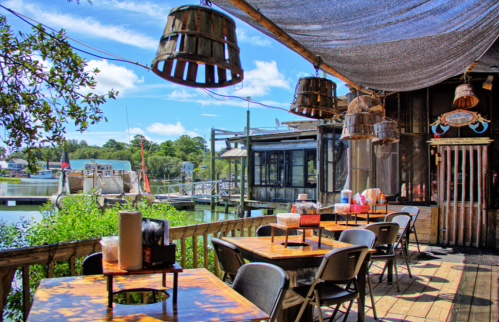 Outdoor dining area with wooden tables, hanging lanterns, and a view of a waterway and greenery under a blue sky.