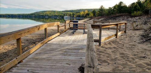 A wooden boardwalk leads to a calm beach with sandy shores and trees in the background under a cloudy sky.