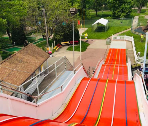 A colorful water slide descends in a park, surrounded by green trees and a mini-golf area in the background.