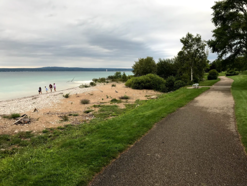 A scenic beach with a walking path, trees, and people strolling along the shore under a cloudy sky.