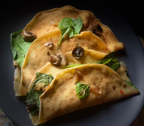 Three savory crepes filled with spinach, mushrooms, and onions, served on a dark plate.