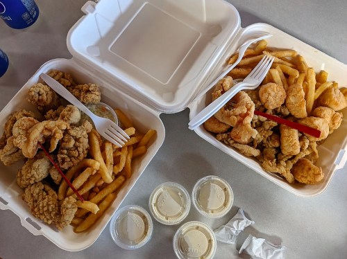 Two takeout containers with fried chicken and fries, accompanied by dipping sauces and plastic utensils on a table.