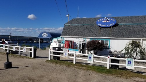 A coastal fishing fleet building with a sign, surrounded by a white fence and a view of the water and boats.