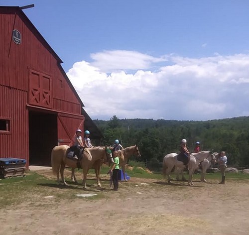 A group of people on horseback near a red barn, surrounded by green trees and a blue sky with clouds.